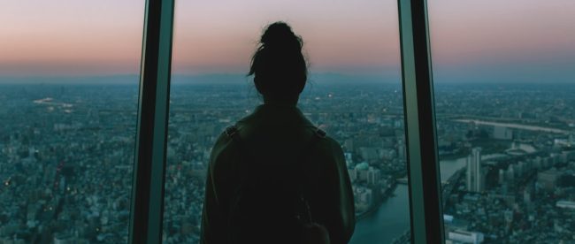 silhouette of person standing inside the building facing the glass mirror watching the city
