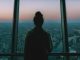 silhouette of person standing inside the building facing the glass mirror watching the city
